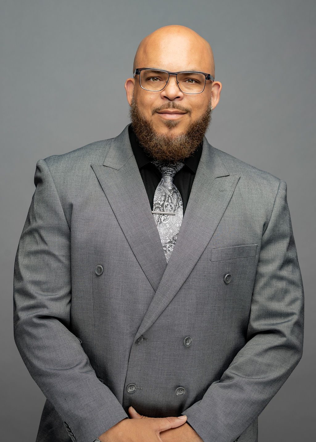 Headshot of Charles "Chuck" Teverbaugh. He is a man with light brown skin and a medium-length dark brown beard. He does not have hair. He is smiling and wearing glasses and a gray suit and tie.