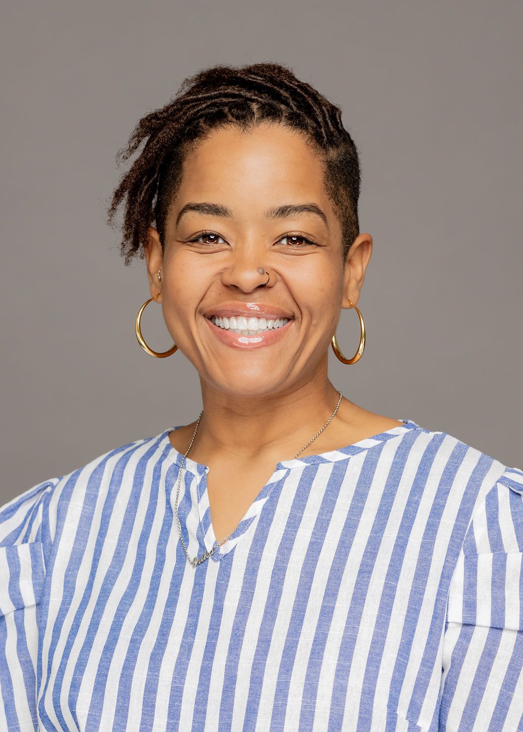 Headshot of Erika Nicole. She is a woman with light-medium brown skin and short dark brown locs with shaved sides. She is smiling and wearing a light blue and white striped blouse.