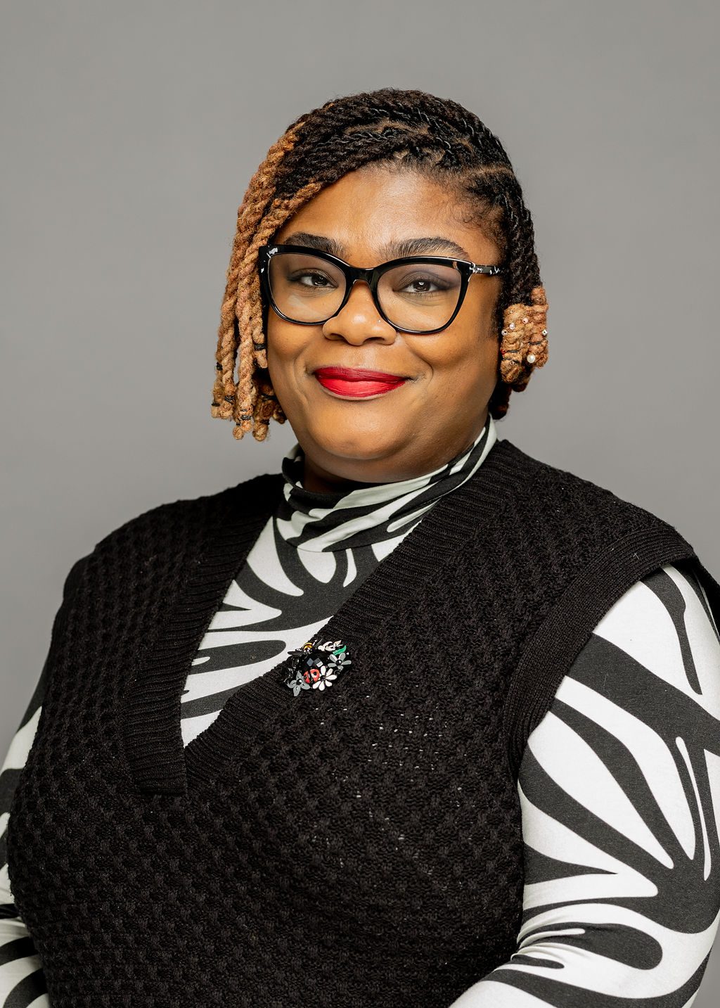 Headshot of Friendella Scott. She is a woman with medium brown skin and chin-length braids that fade from black to light brown. She is smiling and wearing glasses and a black sweater vest over a zebra print button up.