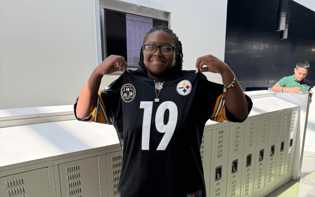 Kahjia Reed, a student at Gary Comer College Prep in Chicago, IL, poses with her favorite sports jersey in the hallway. She is a young woman with dark brown skin, black locs pulled into a low ponytail, and glasses. She is wearing a Steelers jersey and holding it up by the shoulders, smiling.