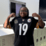 Kahjia Reed, a student at Gary Comer College Prep in Chicago, IL, poses with her favorite sports jersey in the hallway. She is a young woman with dark brown skin, black locs pulled into a low ponytail, and glasses. She is wearing a Steelers jersey and holding it up by the shoulders, smiling.