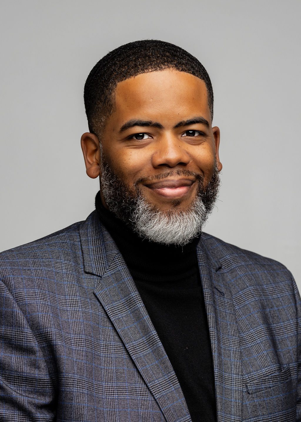 Headshot of Justin Boyd. He is a man with medium-brown skin, black hair shaved into a buzz cut, and a salt and pepper beard. He is smiling and wearing a dark gray blazer over a black turtleneck.