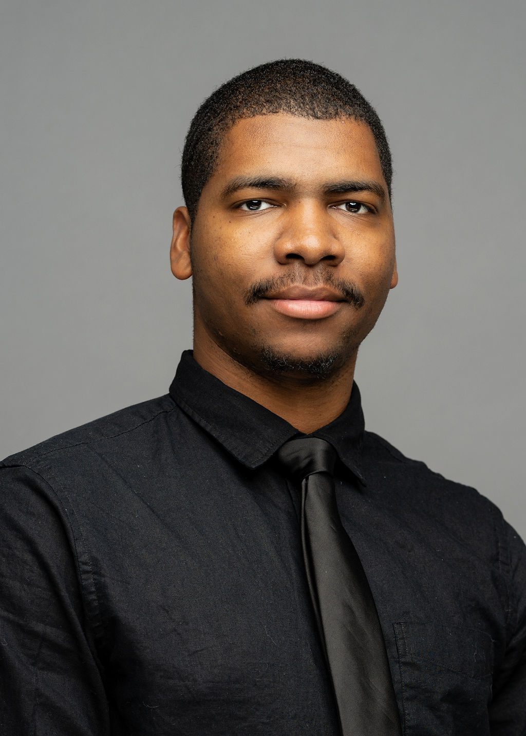 Headshot of Kevin McLurkin. He is a man with medium brown skin and a short black buzz cut and stubble. He is smiling slightly and wearing a black button-up with a thin black tie.