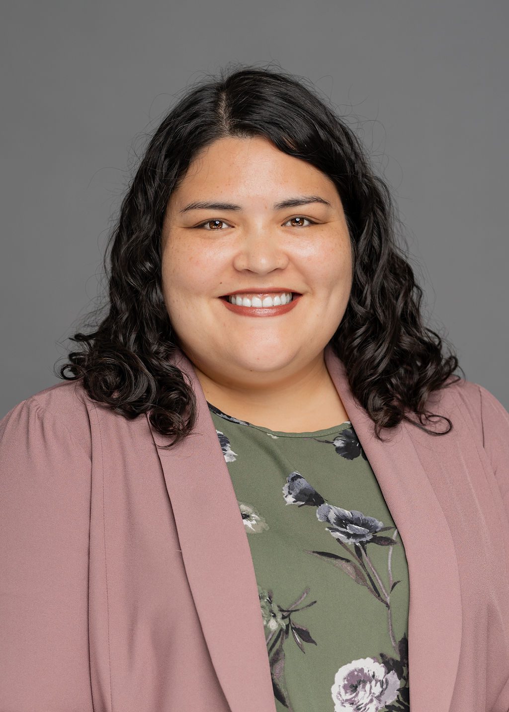 Headshot of Melanie Chwee. She is a woman with light brown skin and wavy black shoulder-length hair. She is smiling and wearing a dusty pink blazer over an olive green floral blouse.