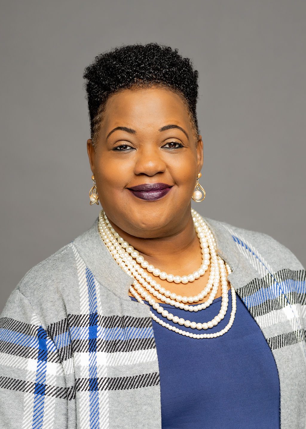 A professional headshot of Ronda Coleman. She is a woman with light brown skin and short black natural hair that's shaved on the sides. She is smiling and wearing a gray, blue, and white cardigan on top of a blue blouse. A set of pearl necklaces lays around her neck.