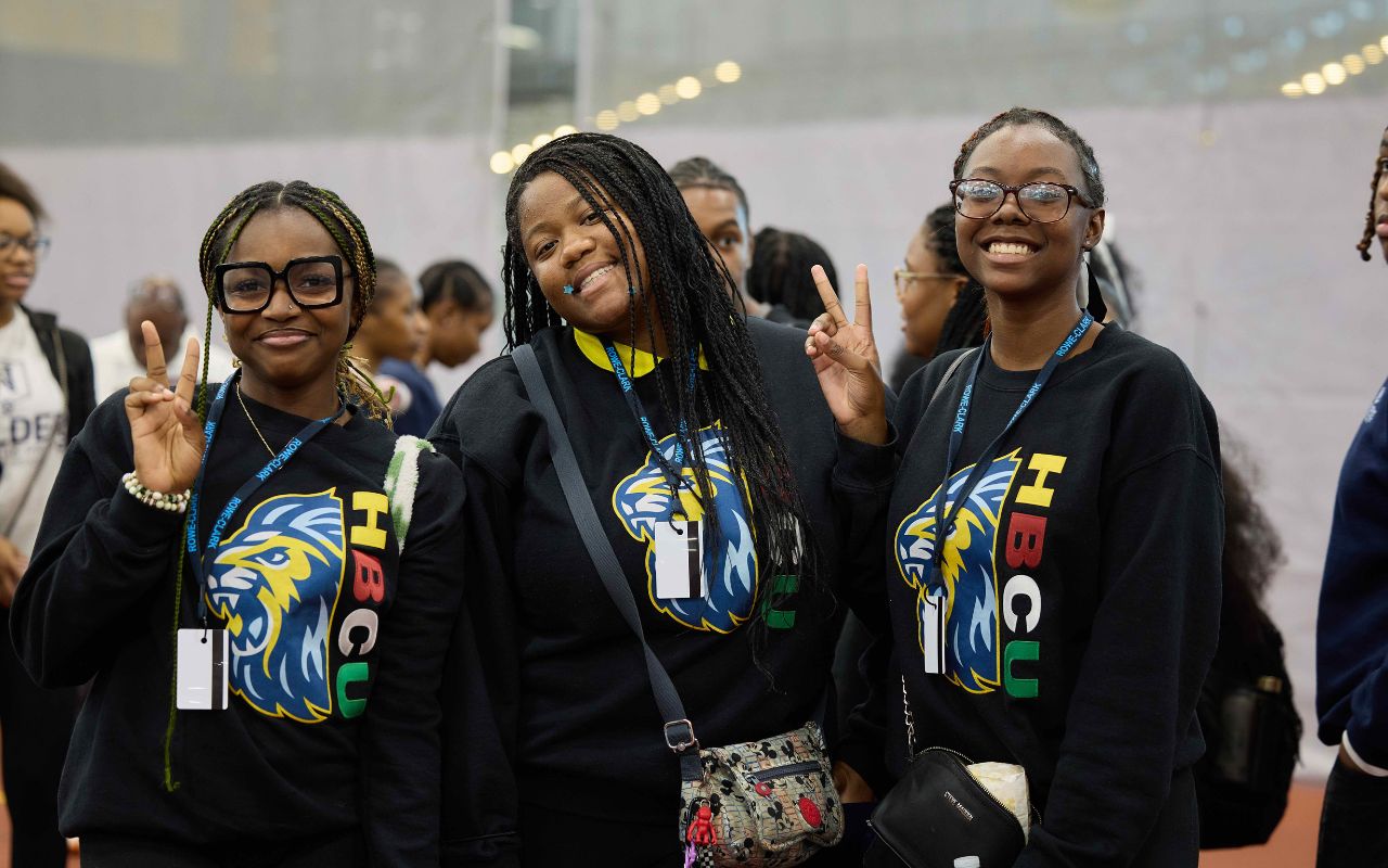 Three Rowe-Clark Math & Science Academy students smile and pose at the HBCU College Fair. They are all wearing matching black sweatshirts with the Rowe-Clark mascot next to the letters HBCU.