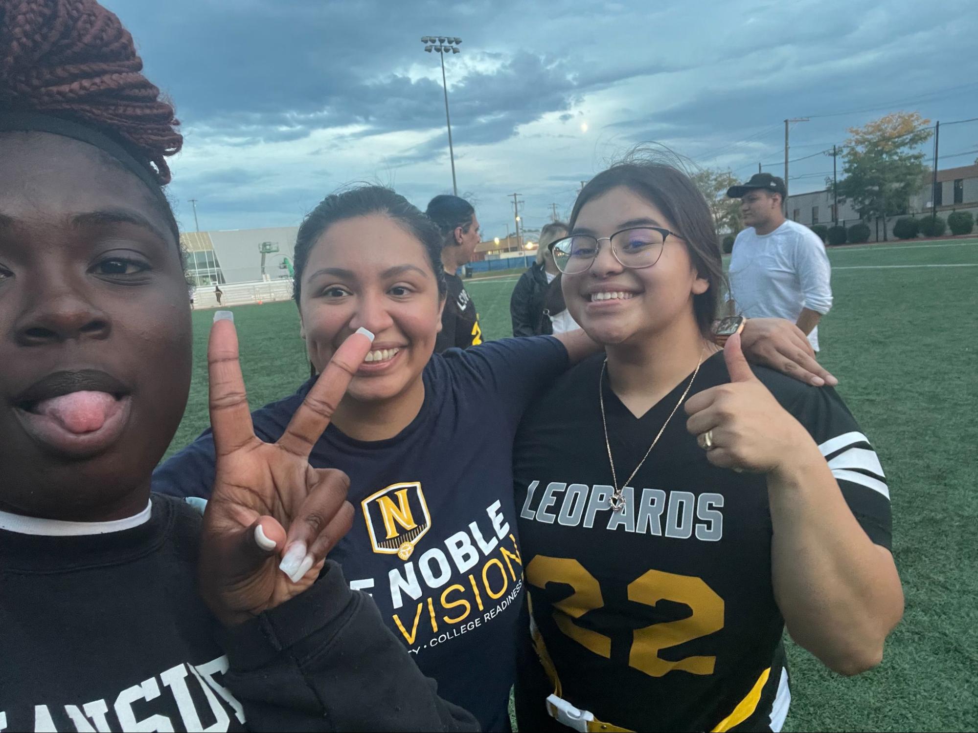 Sanchez taking a selfie with two of her students in front of a football field. The student on the left in the front is posing with the peace sign and the one on Sanchez's right is giving a thumbs up. Sanchez smiles wide between the two of them, her arm over the shoulder of the student on her right.