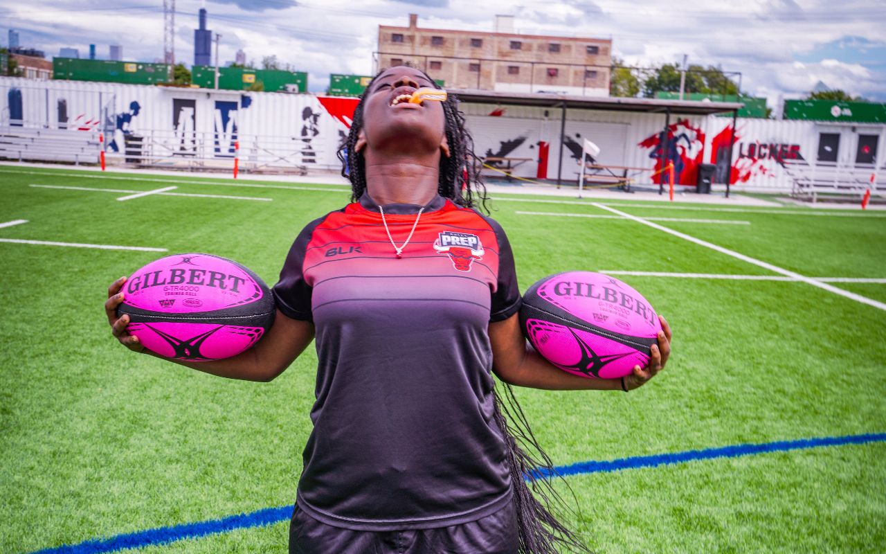 Amira is standing triumphantly, her head leaning back as though to yell to the sky as she holds rugby balls in both hands. She grips a mouthguard between her teeth. Amira is a young woman with dark brown skin and long black braids that go past her waist. She is wearing a black and red Bulls Prep jersey and is standing outside on a field.