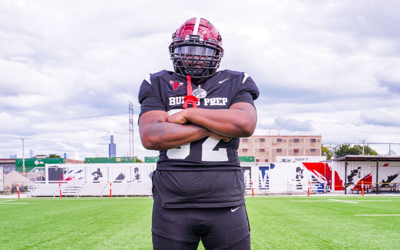 Damari stands in his black and red football helmet and Bulls Prep jersey on a field. He is crossing his arms and looking confidently at the camera. He is a young man with dark brown skin.