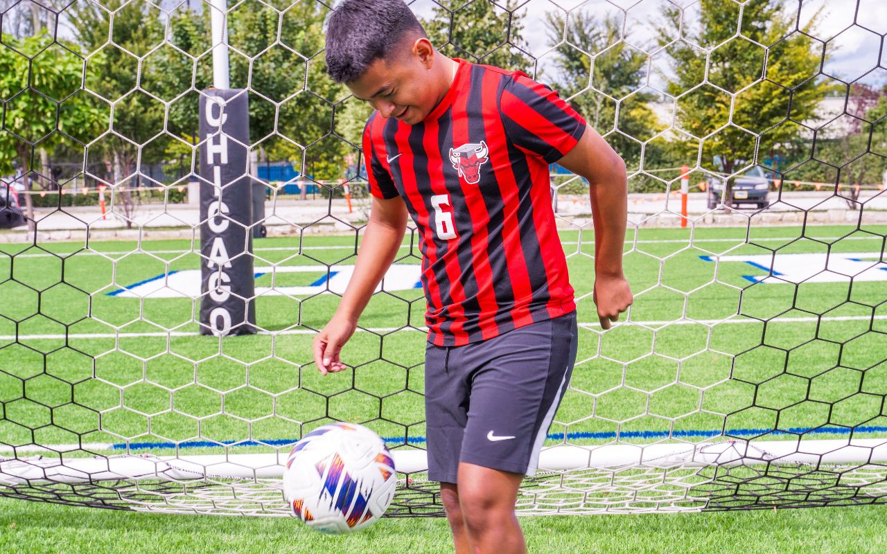 Dida is looking down, smiling, as he kicks up a soccer ball. He is wearing a red and black striped Bulls Prep soccer jersey and outside on a field. Dida is a young man with light brown skin and short brown hair that is shaved on the sides.