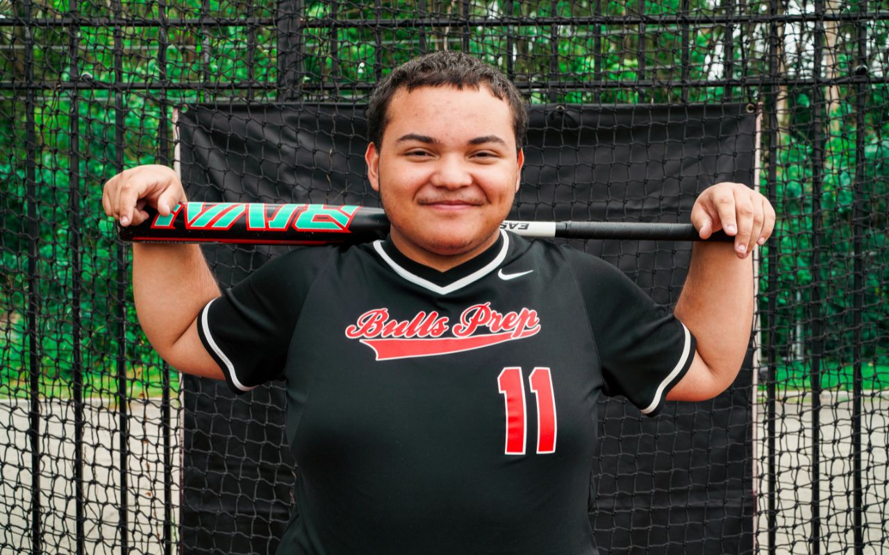 Headshot of Isaiah. He is smiling and holds a softball bat behind his shoulders. He's wearing a black, red, and white Bulls Prep jersey with the number 11 on it. Isaiah is a young man with light brown skin and very short brown hair.