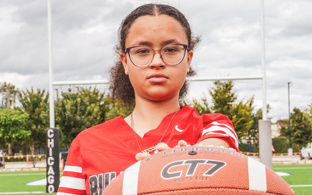 Headshot of Lilian. She is on a football field and wearing a red and white Bulls Prep jersey and holding a football out in front of her. Her face is stoic. She is a young woman with light brown skin, short curly brown hair tied into a small ponytail, and glasses.