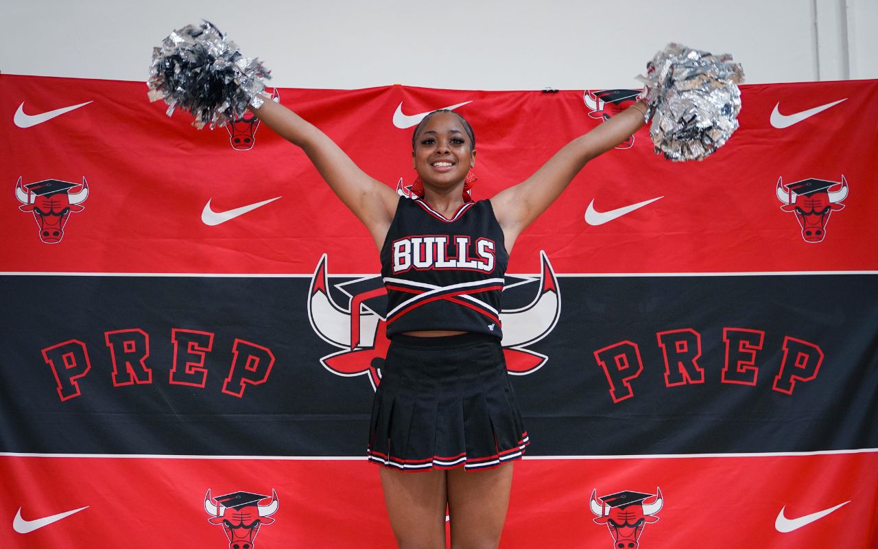 Armani is standing with her arms outstretched above her, holding pom poms in each hand. She is smiling wide and wearing a black and red Bulls Prep cheerleading outfit. Behind her, there is a Bulls Prep photo backdrop with the mascot decal. Armani is a young woman with medium brown skin and medium-length black braids pulled back into a ponytail.