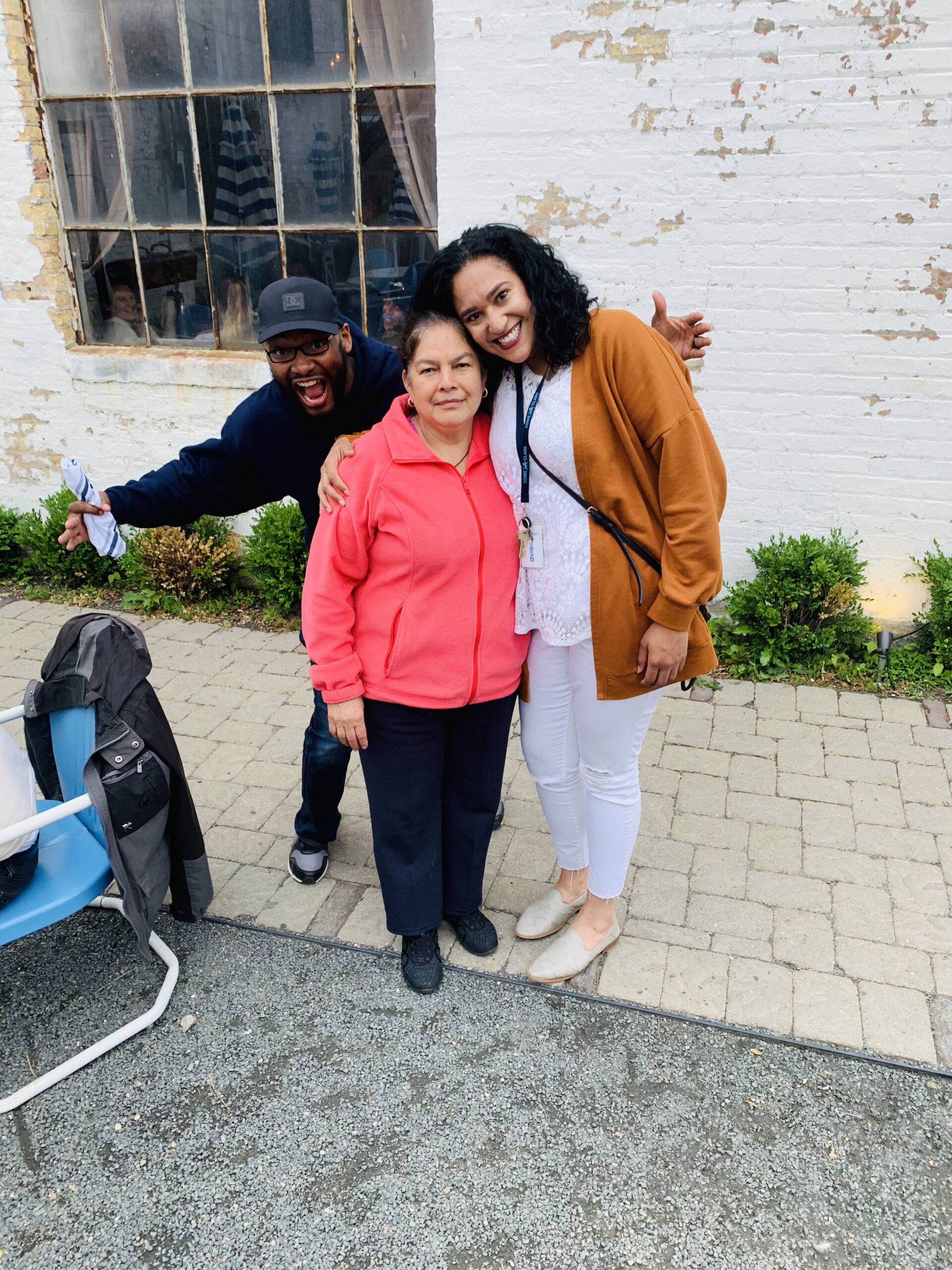 Cora is smiling and side hugging a Rowe-Clark staff member while Burns comes up behind them with his arms outstretched in a funny pose. They are all smiling and in front of a white brick building outside.