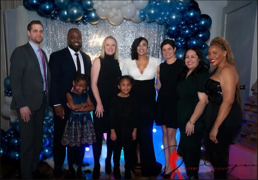 A group of people in formal dress standing in front of a sparkly photo backdrop. Cora is in the center and everyone is smiling.