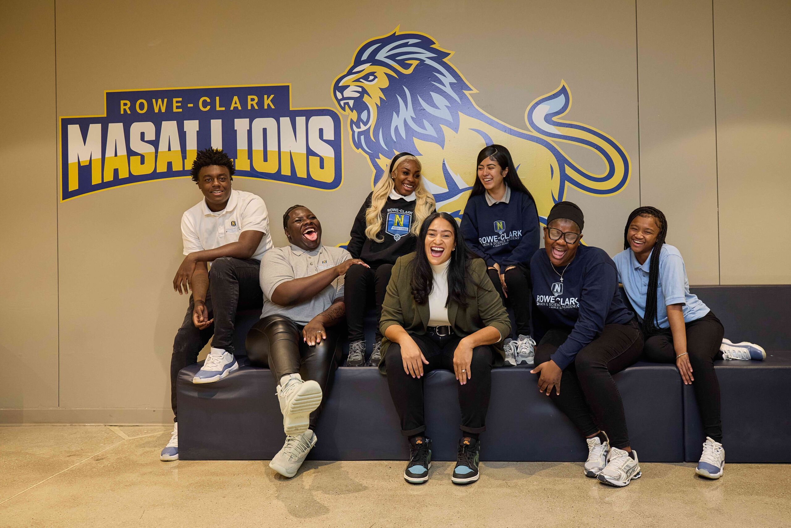 Cora sits on a couch with many Rowe-Clark students sitting around her. They are all laughing and look like they're having a good time. Behind them, you can see a giant wall decal of the Rowe-Clark Masai Lions mascot and logo.