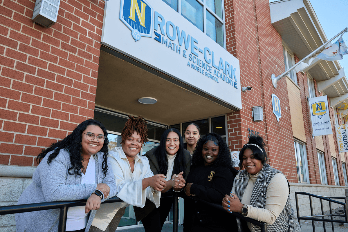 Cora and several Rowe-Clark staff members lean on a railing in front of the school's entrance. They are all looking at the camera and smiling.