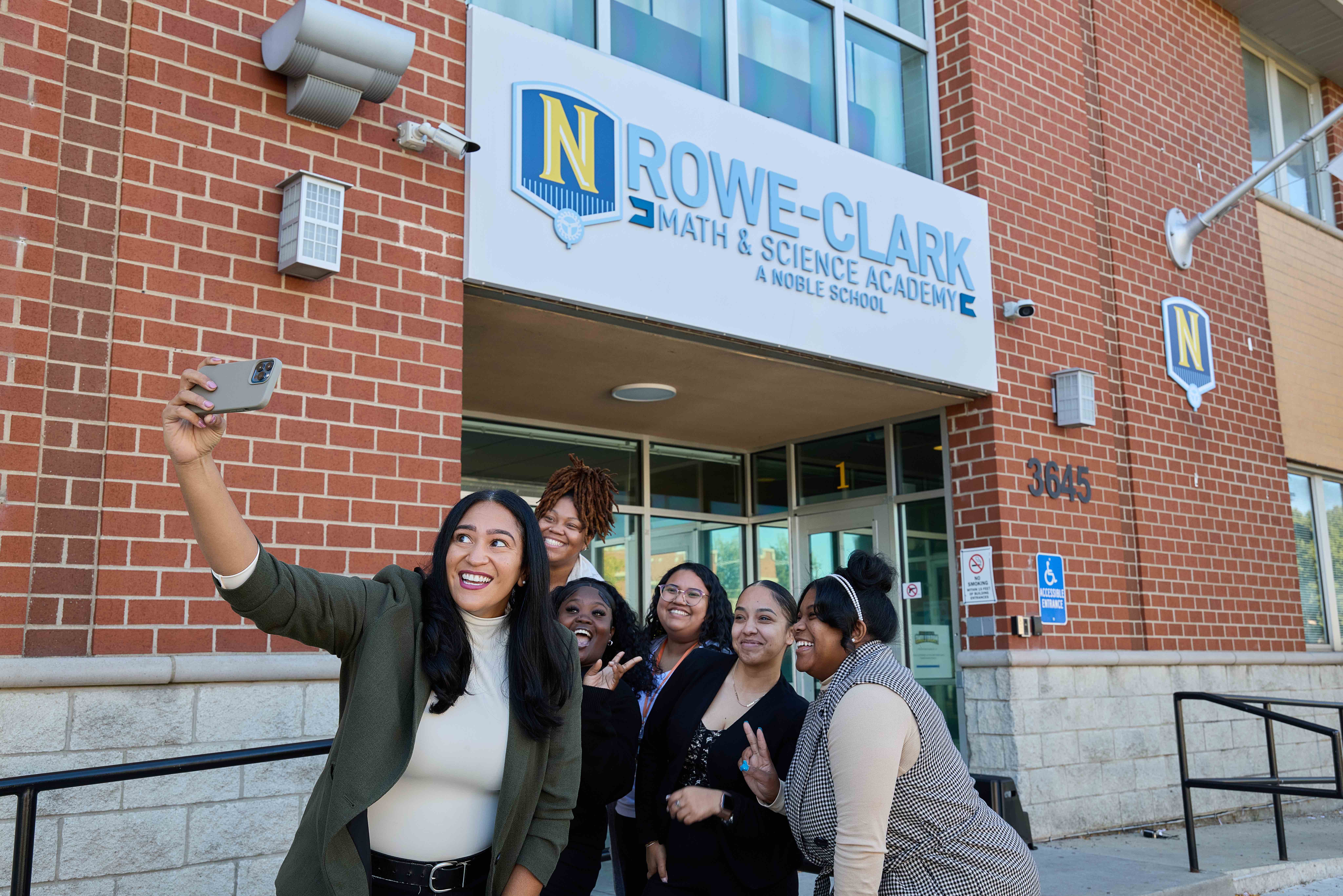 Cora holds a phone in her hands to take a selfie as several of her former students gather behind her to pose. They are in front of the Rowe-Clark Math & Science Academy's entrance.