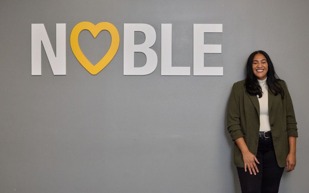 Brenda Cora, Noble Schools' new CEO, stands smiling next to a large white wall decal that says "Noble" with a yellow heart taking the place of the O. Cora is a woman with a medium-light skin tone and medium-length straight black hair. She is wearing a gray blazer over a white turtleneck and black jeans.