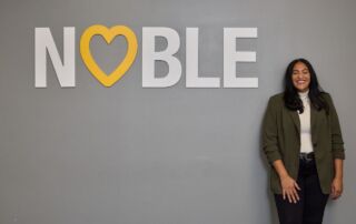 Brenda Cora, Noble Schools' new CEO, stands smiling next to a large white wall decal that says "Noble" with a yellow heart taking the place of the O. Cora is a woman with a medium-light skin tone and medium-length straight black hair. She is wearing a gray blazer over a white turtleneck and black jeans.