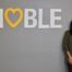 Brenda Cora, Noble Schools' new CEO, stands smiling next to a large white wall decal that says "Noble" with a yellow heart taking the place of the O. Cora is a woman with a medium-light skin tone and medium-length straight black hair. She is wearing a gray blazer over a white turtleneck and black jeans.