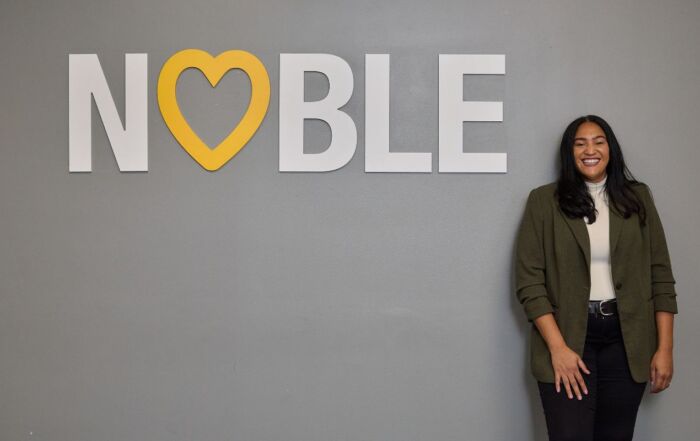 Brenda Cora, Noble Schools' new CEO, stands smiling next to a large white wall decal that says "Noble" with a yellow heart taking the place of the O. Cora is a woman with a medium-light skin tone and medium-length straight black hair. She is wearing a gray blazer over a white turtleneck and black jeans.