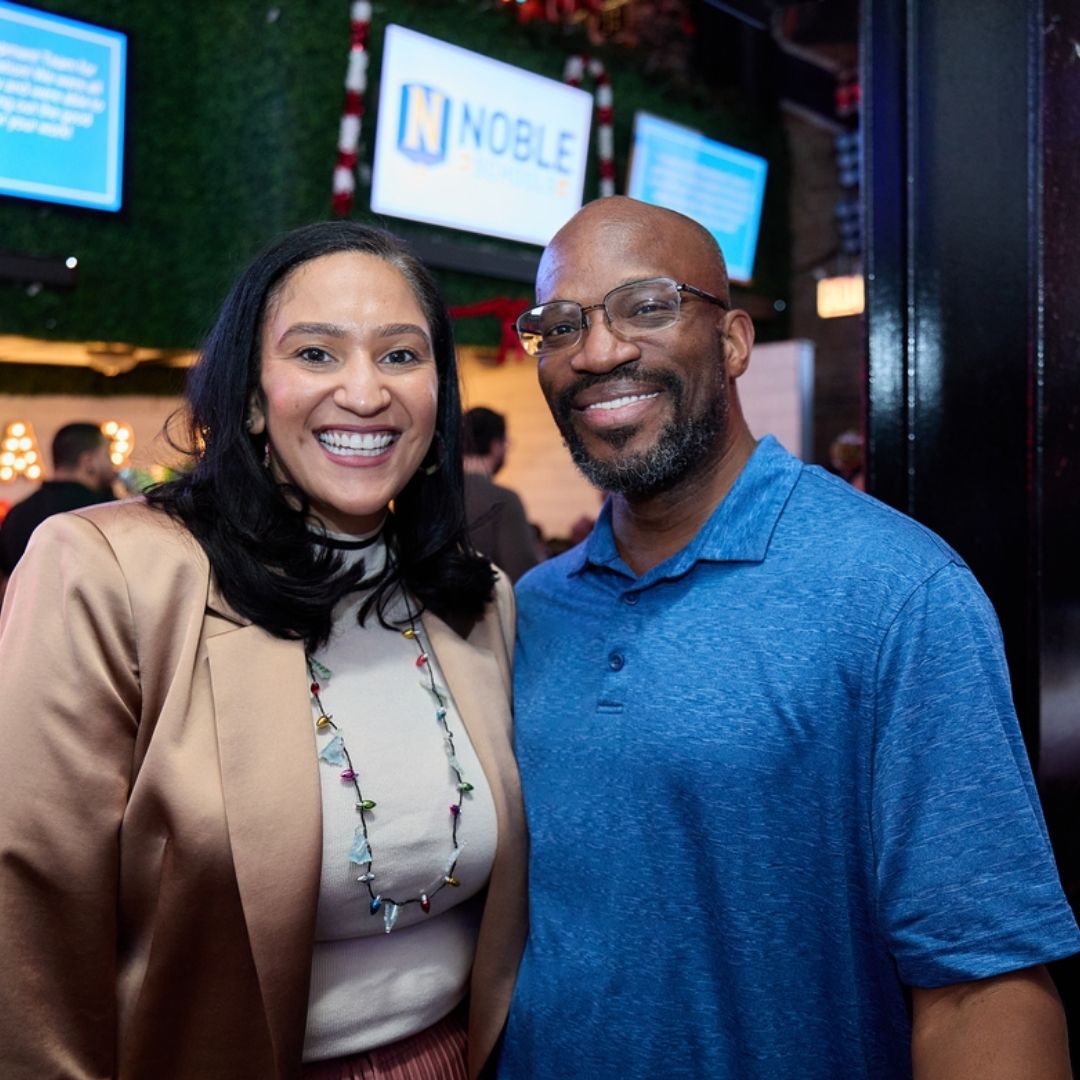 Cora stands next to her husband Donald. They are smiling in front of the scene of a Noble staff party.