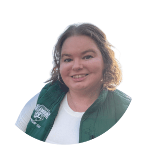 A headshot of Amy Agosta. She is a woman with a light skin tone and short curly blonde hair. She is wearing a white long-sleeved shirt underneath a puffy green Gary Comer College Prep vest with the mascot on it. The background is cut out.