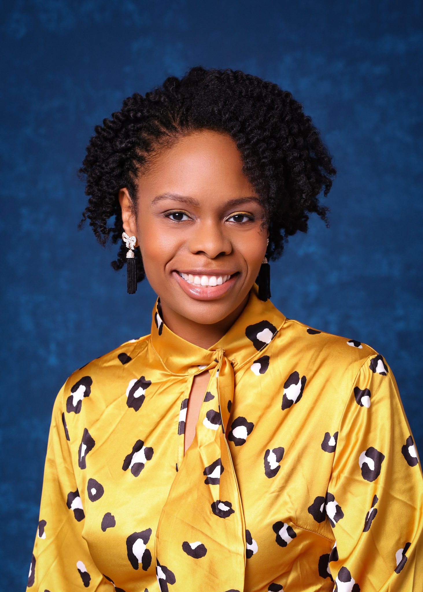 A headshot of Pattilyn Beals. She is a woman with a medium-dark brown skin tone and short black natural hair in coils. She is wearing a bright gold silk blouse with a leopard pattern on it and is standing in front of a dark blue background.