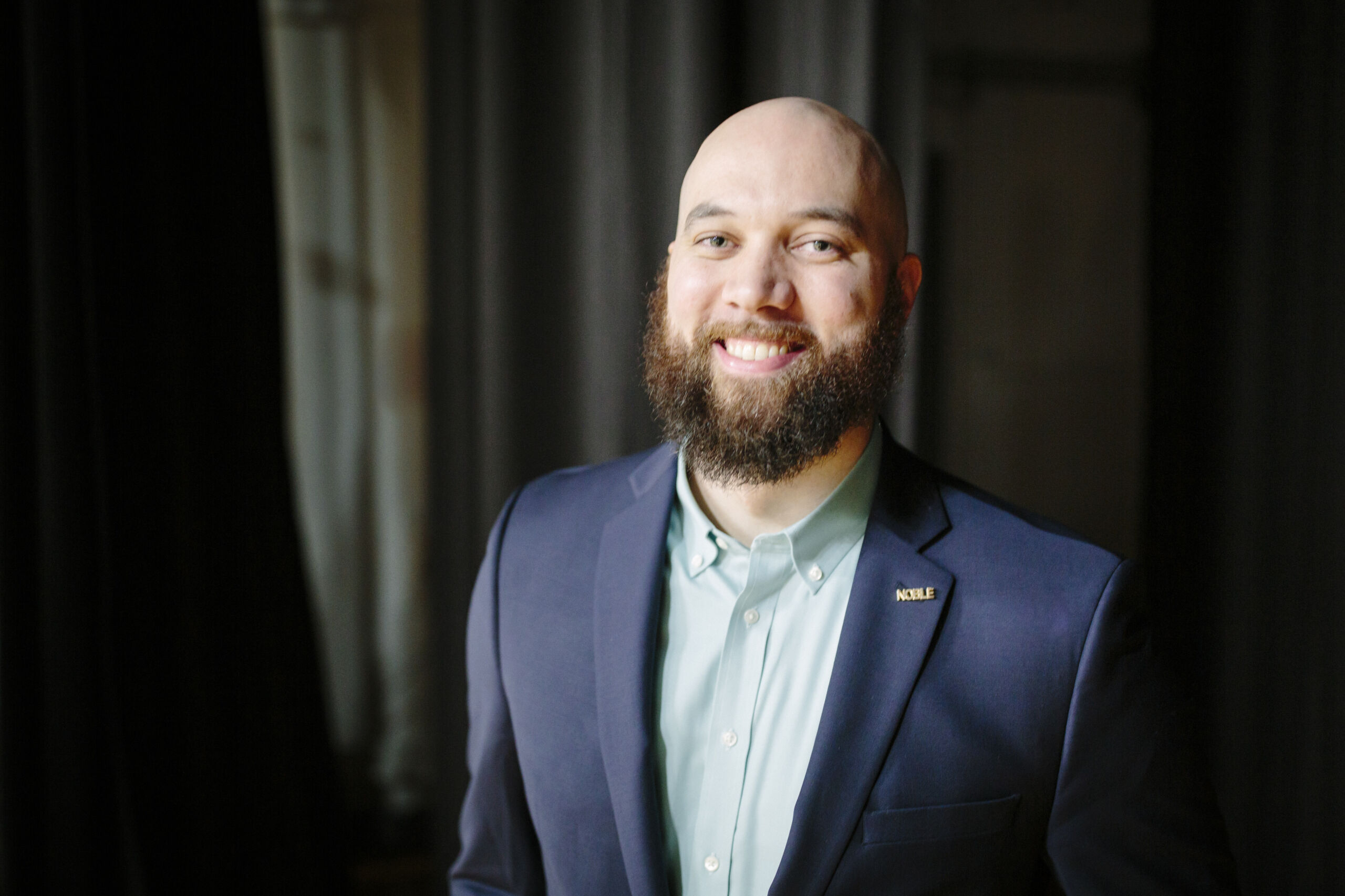 A headshot of David Brown. He is a Black man with a light skin tone and a full brown beard. He is wearing a light blue button-up underneath a dark blue blazer with a gold pin on it that says "Noble". He is standing in front of an indistinct dark background.