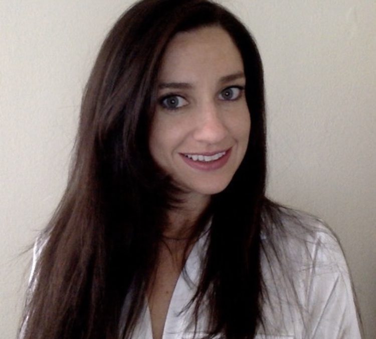 A headshot of Meghan Wagner. She is a woman with a light skin tone and long straight dark brown hair. She is wearing a white blouse and standing in front of a white wall.