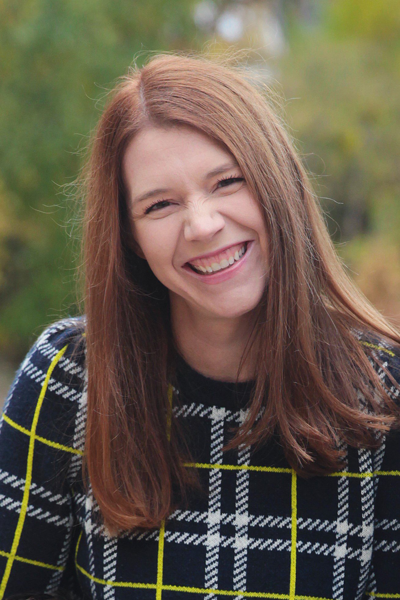 A headshot of Erin Greenfield. She is a woman with a light skin tone and long straight red hair. She is wearing a black, yellow, and white plaid sweater and standing in front of a blurred nature background.