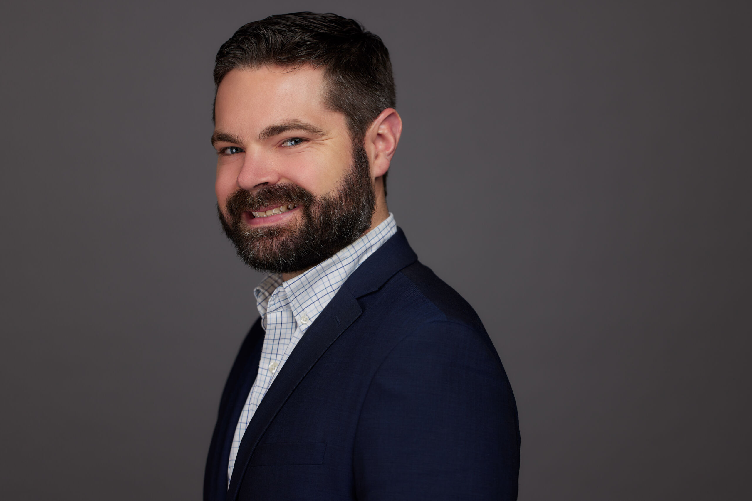 A headshot of R.C. Hyatt. He is a man with a light skin tone and short dark brown hair and beard. He is wearing a graph paper pattern button-up underneath a black blazer. He is standing in front of a blank gray background.