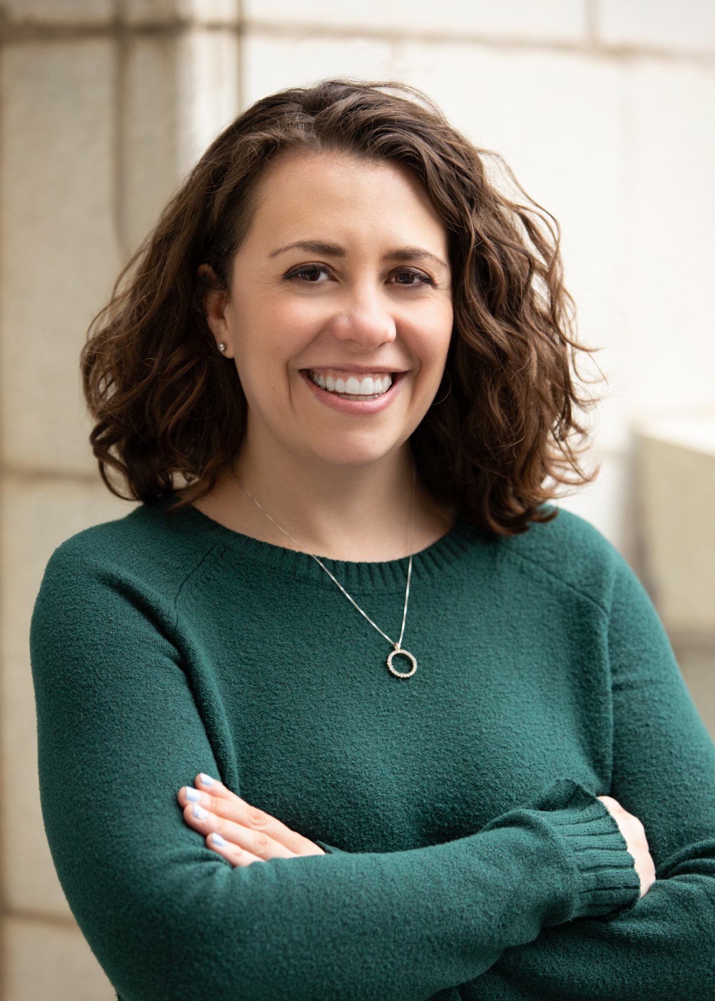 A headshot of Ellen Moiani. She is a woman with a light skin tone and short wavy brown hair. She is wearing a dark green sweater and standing in front of a blurred outdoor background.