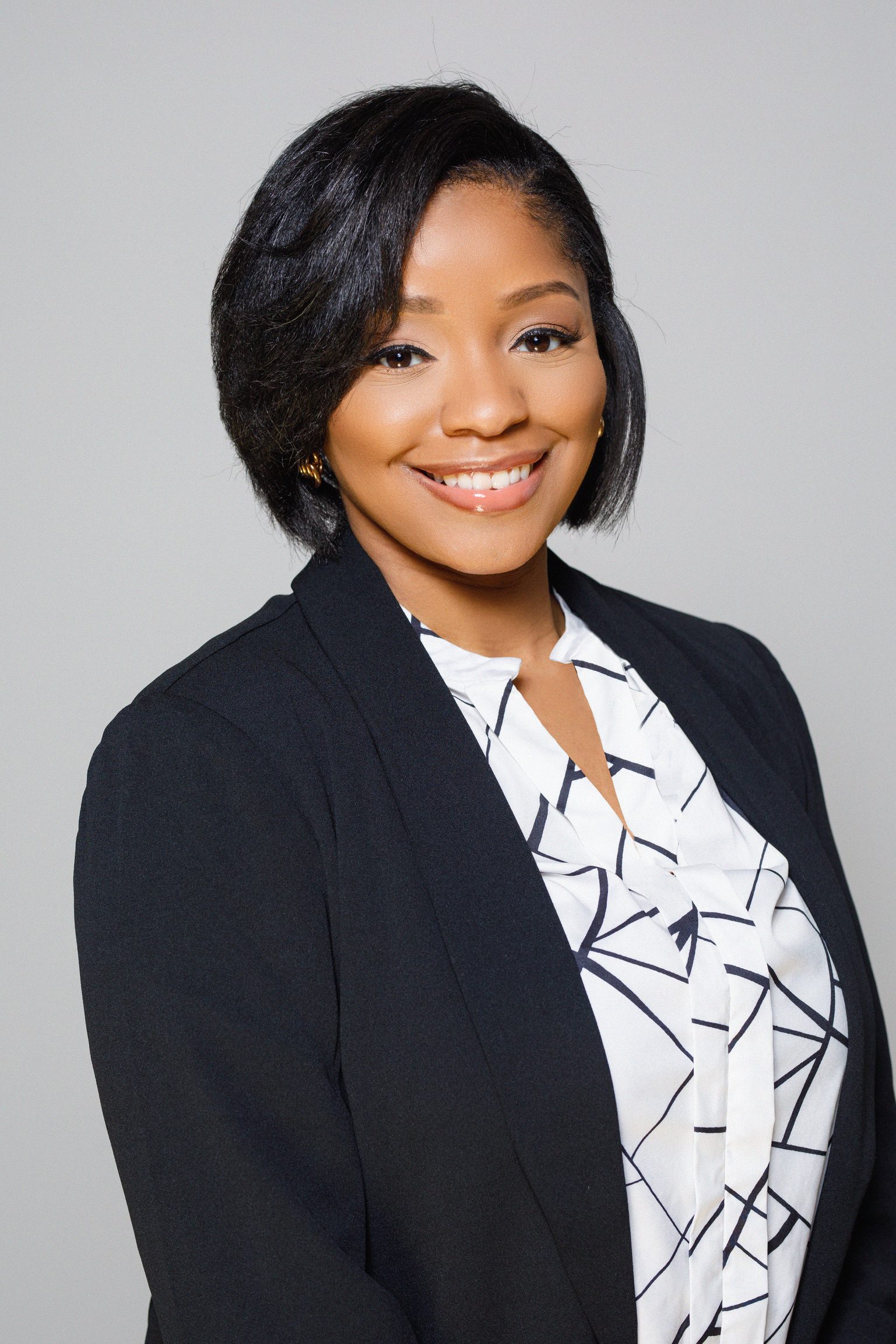 A headshot of Carissa Moorehead. She is a woman with a medium brown skin tone and short straight black hair. She is wearing a white blouse with an abstract black pattern on it underneath a black blazer. She is standing in front of a blank white background.