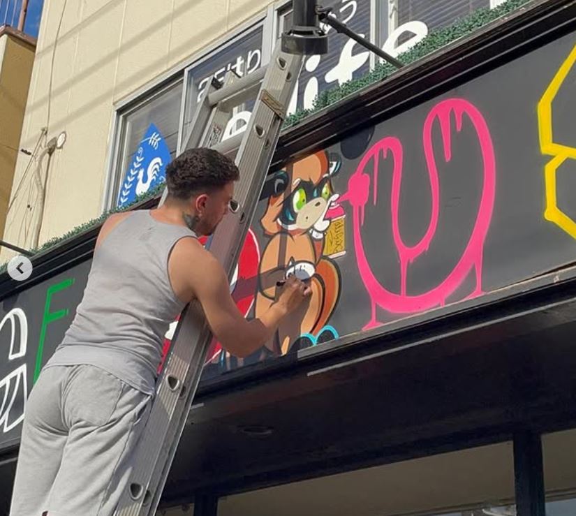 Locander is on a ladder in front of a store sign, painting Richie the Tanuki.