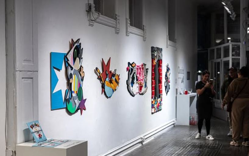 Locander's artwork displayed on a gallery wall. They are brightly painted cutouts of his characters in different layouts.