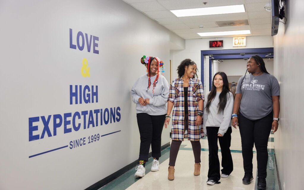 Four students and staff members walk down a hallway at Noble Street College Prep, smiling and talking. On the wall, there is a decal that says "Love and High Expectations since 1999"