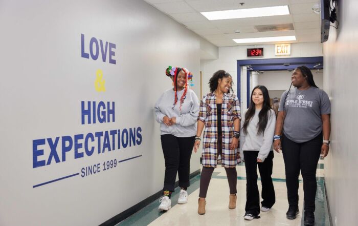 Four students and staff members walk down a hallway at Noble Street College Prep, smiling and talking. On the wall, there is a decal that says "Love and High Expectations since 1999"