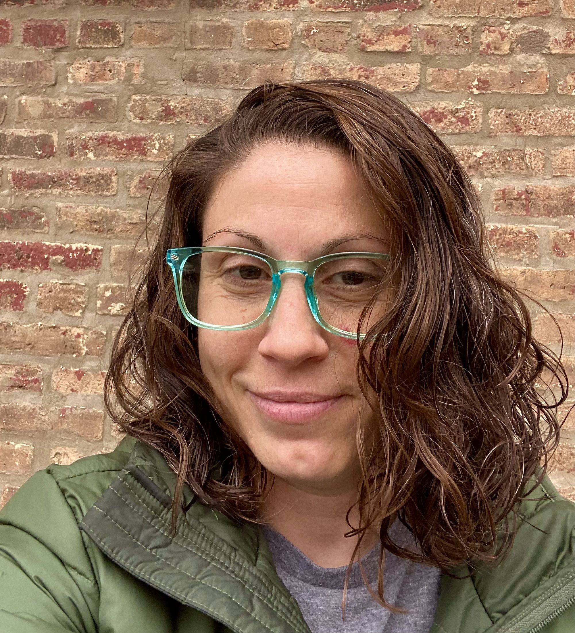 A headshot of Bianca Severino. She is a woman with a light skin tone and medium-length, wavy red hair. She is wearing glasses with light teal frames and a green jacket over a gray t-shirt. Behind her is a brick wall.