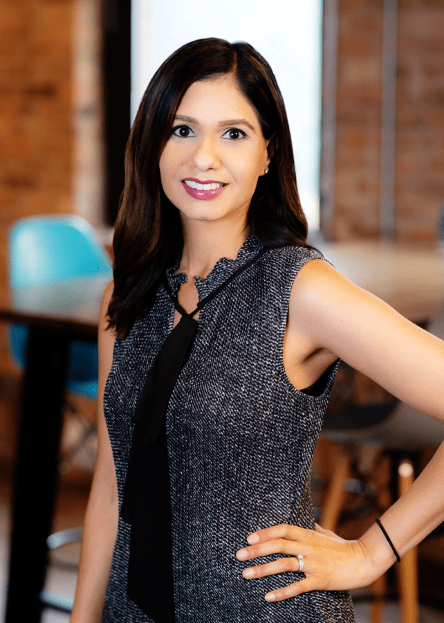 A headshot of Nishi Shah. She is a woman with a light brown skin tone and long dark brown straight hair. She is wearing a black and white sleeveless dress and standing in front of a blurred office background.