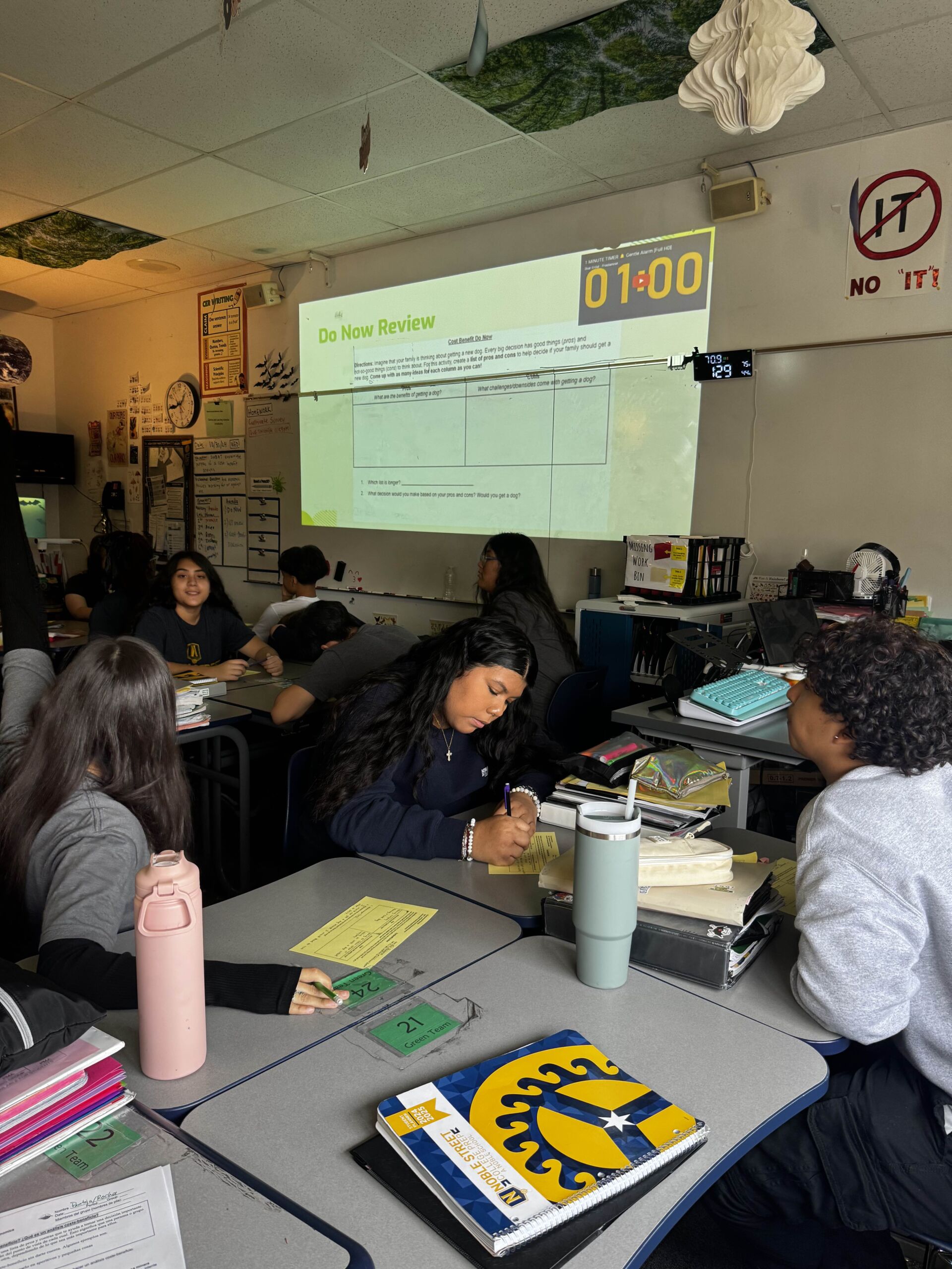 Allison sits at a classroom desk, surrounded by her peers. She is writing on a small slip of paper. Behind her, you can see a projected slide on the white board that has the title "Do Now Review".