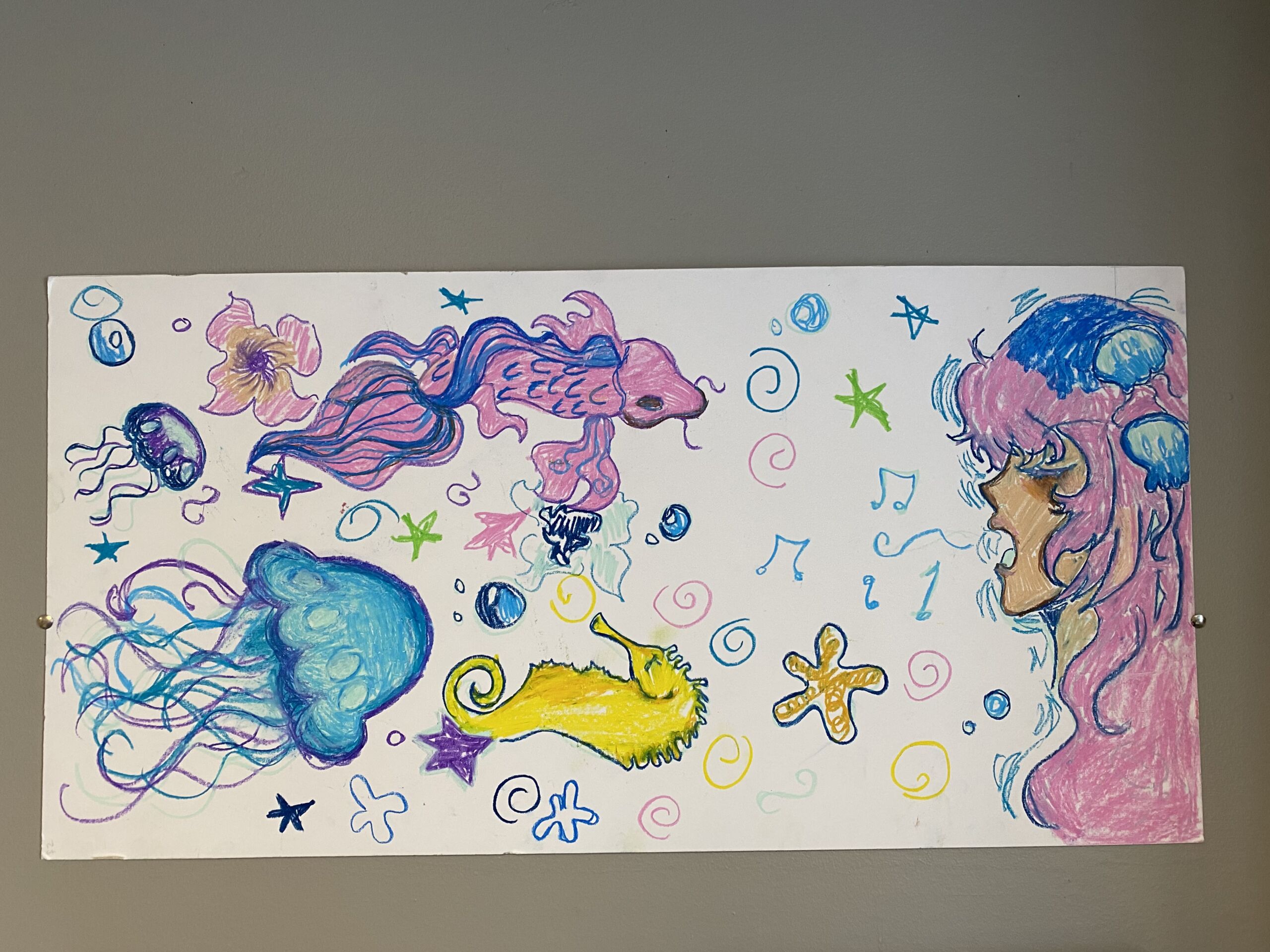 Pastel drawing by Tahini Hargrow-Phelps. It is on a long horizontal piece of white paper and depicts a pink-haired mermaid singing to a group of sea creatures, including a fish, a seahorse, and a jellyfish.