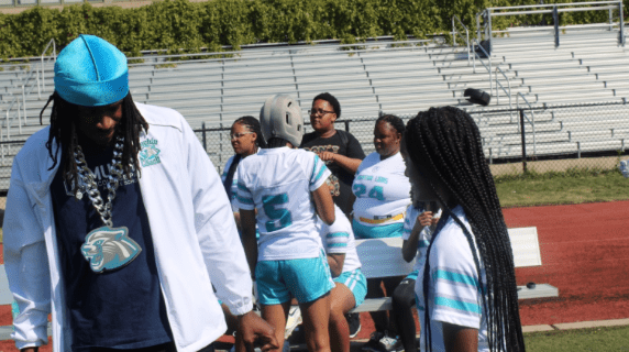 Nick Davis stands, looking down and chatting with a student-athlete. He is wearing a teal durag and white and teal Muchin jacket with the Muchin mountain lion mascot logo on a chain around his neck. You can see other team members chatting in a group behind them on the field.