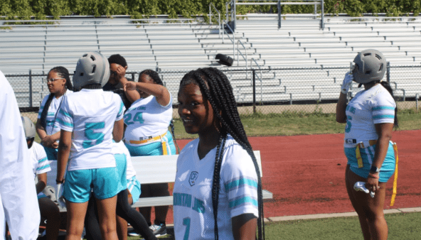 Jauccaria Solomon Jaiccaria Solomon smiles shyly at the camera in her white and teal Muchin jersey. Behind her, you can see her teammates chatting in a group on the football field.
