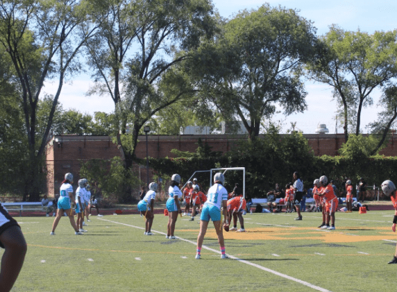 The Muchin team in white and teal uniforms stand in a kickoff line with the Butler team in bright orange uniforms on the football field.