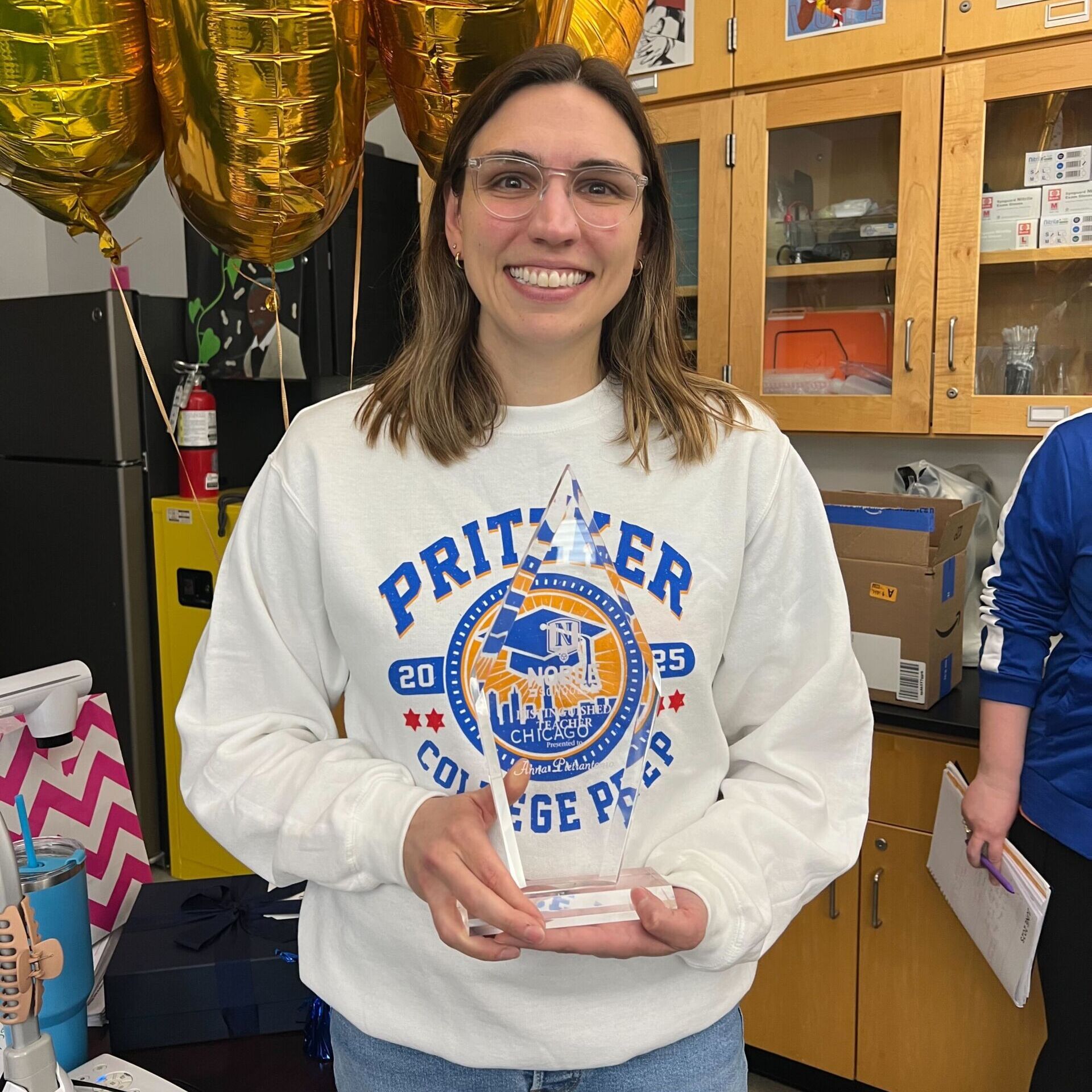 Anna Pietrantonio Anna Pietrantonio, a teacher at Pritzker College Prep in Chicago, is smiling and posing for a photo in her classroom. She is holding a triangular crystal award that has the Noble Schools logo on it and says "Distinguished Teacher" with her name on it. She is a woman with medium-length light brown hair and light skin. She is wearing a white, blue, and orange Pritzker sweatshirt. Behind her, you can see balloons and science equipment in cabinets.