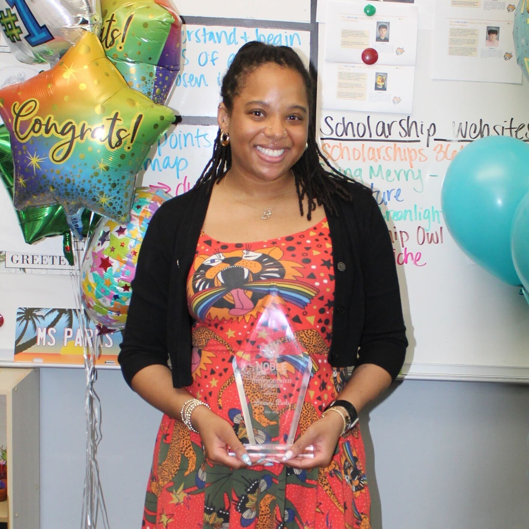 Brittany Parks Brittany Parks, a teacher at Rauner College Prep, stands and smiles for a photo in her classroom. She is holding a triangular crystal award that has the Noble Schools logo on it and says "Distinguished Teacher" with her name on it. She is a woman with shoulder length locs in a ponytail and medium light brown skin. She is wearing a bright, light red dress with a fun pattern of big cats and stars on it. She is standing in front of a whiteboard and some balloons.