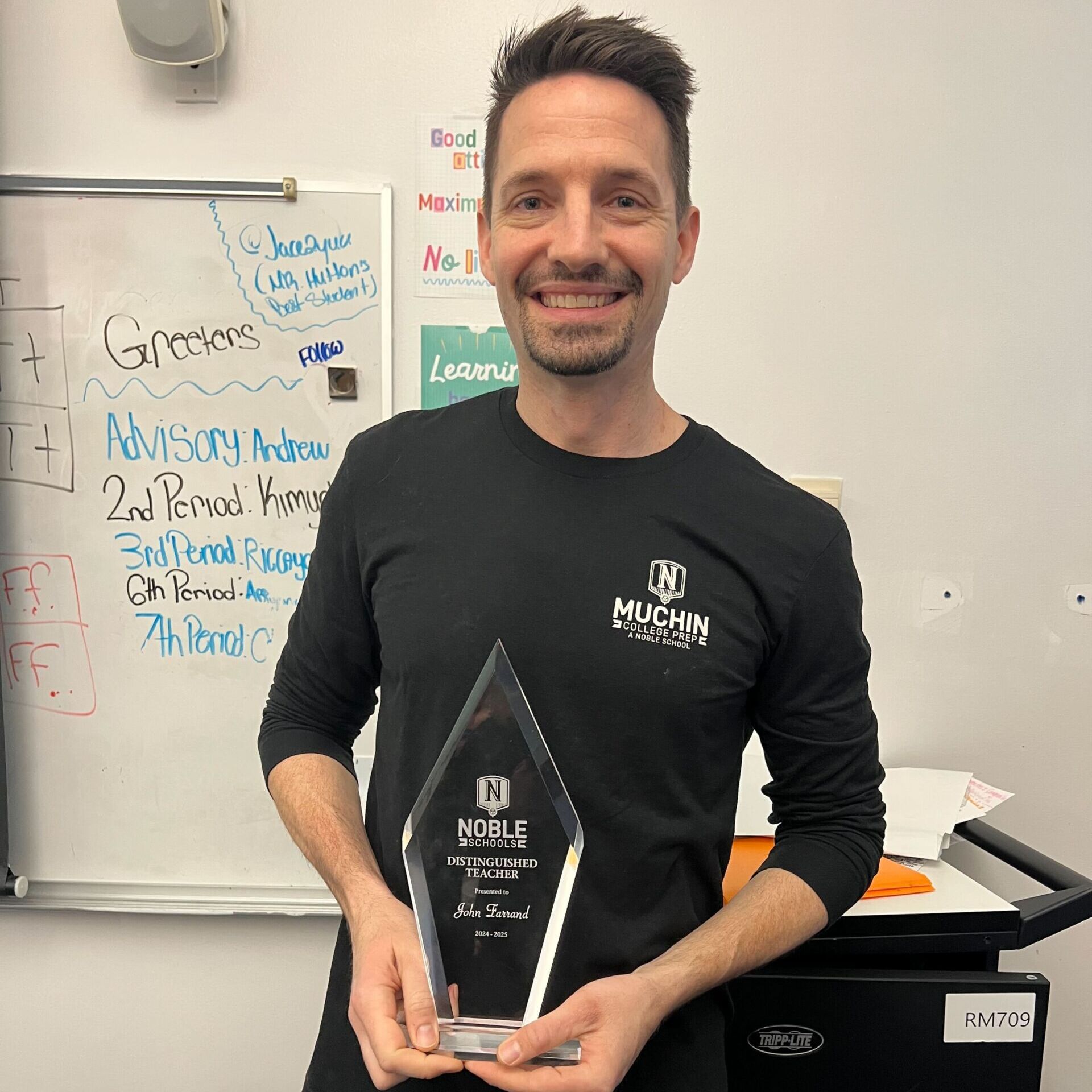 John Farrand John Farrand, a teacher at Muchin College Prep in Chicago, smiles and poses with a triangular crystal award that says "Distinguished Teacher" with his name. He stands in front of a whiteboard. He is a man with short brown hair, a light beard, and light skin. He is wearing a black Muchin sweatshirt.