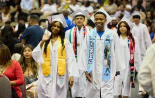 UIC College Prep graduates walk down the aisle in white graduation robes. They are smiling.